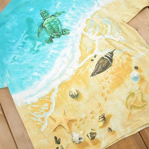 Puede incluir: Una camiseta tie-dye con una escena de playa que presenta una tortuga marina nadando en el oc&eacute;ano, un p&aacute;jaro en la arena y conchas marinas. La camiseta es de color azul claro y amarillo.