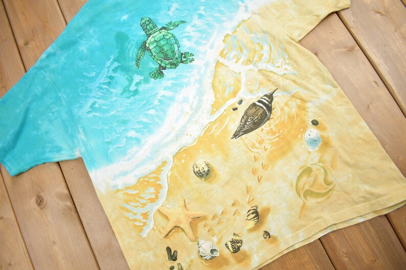 Puede incluir: Una camiseta tie-dye con una escena de playa que presenta una tortuga marina nadando en el oc&eacute;ano, un p&aacute;jaro en la arena y conchas marinas. La camiseta es de color azul claro y amarillo.