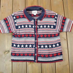 Può includere: Un cardigan di maglia a maniche corte con chiusura a bottoni. Il cardigan è bianco con strisce rosse e blu e presenta un motivo di stelle e cuori.