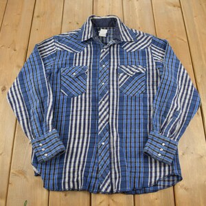 Può includere: Una camicia a maniche lunghe a quadri blu e bianchi con colletto abbottonato e due tasche sul petto.