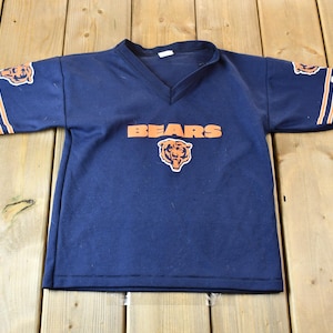 Pode incluir: Uma camiseta azul marinho com listras laranja e brancas nas mangas. A camiseta tem a palavra "BEARS" impressa na frente em letras laranja, com um logotipo de cabeça de urso embaixo.