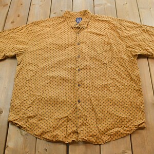 Könnte beinhalten: Ein senfgelbes Button-Down-Hemd mit einem sich wiederholenden Muster aus kleinen, dunkelblauen Formen. Das Hemd hat kurze Ärmel, einen Kragen und eine Brusttasche. Das Gap-Logo ist auf dem Kragen sichtbar.