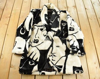 Peut inclure: Un manteau en fausse fourrure noir et blanc avec un motif répété de visages stylisés. Le manteau a un col châle et des manches longues. Le design est audacieux et graphique, avec les visages en noir sur un fond crème.