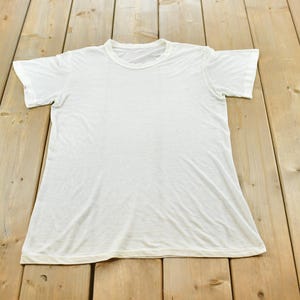 Könnte beinhalten: Ein schlichtes weißes T-Shirt mit kurzen Ärmeln und einem Rundhalsausschnitt. Das Kleidungsstück ist aus einem leichten Stoff gefertigt und verfügt über eine Standardpassform mit gesäumten Ärmeln und einem geraden Saum.