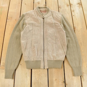 May include: A tan suede jacket with a ribbed knit collar, cuffs, and sleeves. The jacket has a zip-up front and a tailored fit. The sleeves are a lighter tan color than the suede front. The jacket is laid flat on a wooden surface.