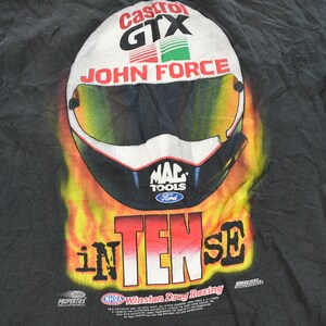 Op de afbeelding: Zwart T-shirt met een racehelm-ontwerp, met de woorden "Castrol GTX" en "John Force". De helm staat tegen een achtergrond van vlammen, met "INTENSE" eronder. Inclusief logo's van "MAC TOOLS" en "Ford".