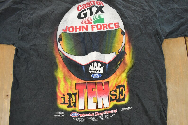 Op de afbeelding: Zwart T-shirt met een racehelm-ontwerp, met de woorden "Castrol GTX" en "John Force". De helm staat tegen een achtergrond van vlammen, met "INTENSE" eronder. Inclusief logo's van "MAC TOOLS" en "Ford".