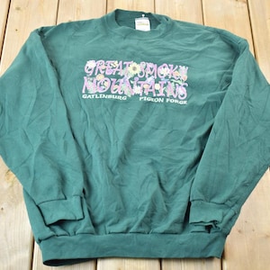 May include: Green long-sleeved sweatshirt with the text "Great Smoky Mountains" in pink and purple lettering with floral accents. The text "Gatlinburg Pigeon Forge" is printed below the main text.