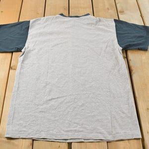 May include: A gray t-shirt with dark blue sleeves and collar. The shirt is laid flat on a wooden surface, showcasing its design. The t-shirt is a casual wear item.