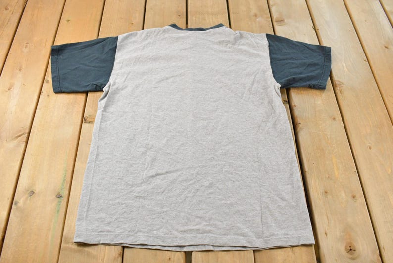 May include: A gray t-shirt with dark blue sleeves and collar. The shirt is laid flat on a wooden surface, showcasing its design. The t-shirt is a casual wear item.