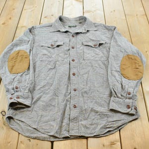 May include: A long-sleeve button-down shirt in a heathered gray fabric featuring a standard collar, two chest pockets with buttoned flaps, and contrasting tan elbow patches. The shirt includes buttoned cuffs and a full-length front button closure.