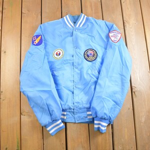 Op de afbeelding: Een lichtblauwe bomberjack met witte en blauwe geribbelde boorden aan de kraag, manchetten en tailleband. De jas heeft drie patches aan de voorkant: een ronde patch met een rood, wit en blauw ontwerp, een ronde patch met een geel en blauw ontwerp en een driehoekige patch met een rood, wit en blauw ontwerp.