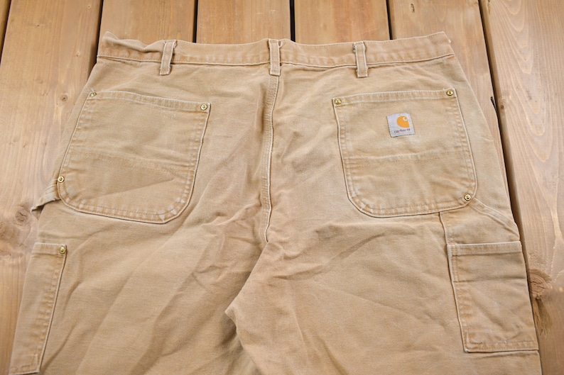 Puede incluir: Un pantal&oacute;n de trabajo de color beige con varios bolsillos, incluido un bolsillo grande en la pierna derecha con un parche de logotipo de Carhartt.