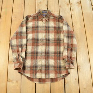May include: A long-sleeved, button-down shirt with a plaid pattern in shades of brown, beige, and black. The shirt has a classic collar and buttoned cuffs. The garment is laid flat on a wooden surface.