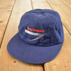 May include: A navy blue baseball cap with the Raytheon logo embroidered on the front. The logo includes the word "Raytheon" in red, "ON BOARD" in white, and a gray swoosh design. The cap is made of a soft fabric.