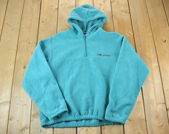 Puede incluir: Una sudadera con capucha de forro polar turquesa con cierre de media cremallera y capucha. La palabra "POLARIS" está impresa en negro en el pecho. El dobladillo inferior es elástico. La sudadera está sobre una superficie de madera.