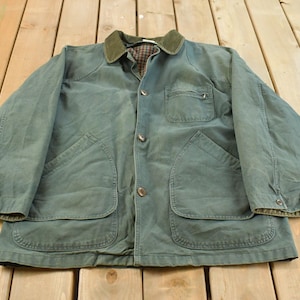 Op de afbeelding: Een vintage groene jas met een corduroy kraag en vier voorzakken. De jas heeft een knoopsluiting en een geruite voering zichtbaar bij de kraag en manchetten. De jas ligt plat op een houten oppervlak.