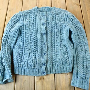 May include: A light blue, button-down cardigan sweater with a round neckline. The sweater features a cable knit design with bobble stitch detailing and is displayed on a wooden surface.