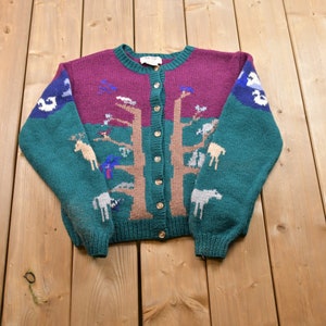 May include: A green and burgundy knitted cardigan with a brown tree and animal designs. The cardigan has a button closure.