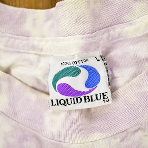 May include: A white and purple tie-dye t-shirt with a Liquid Blue tag. The tag is white with a blue and purple swirl design and the text "100% Cotton" and "Liquid Blue TM".