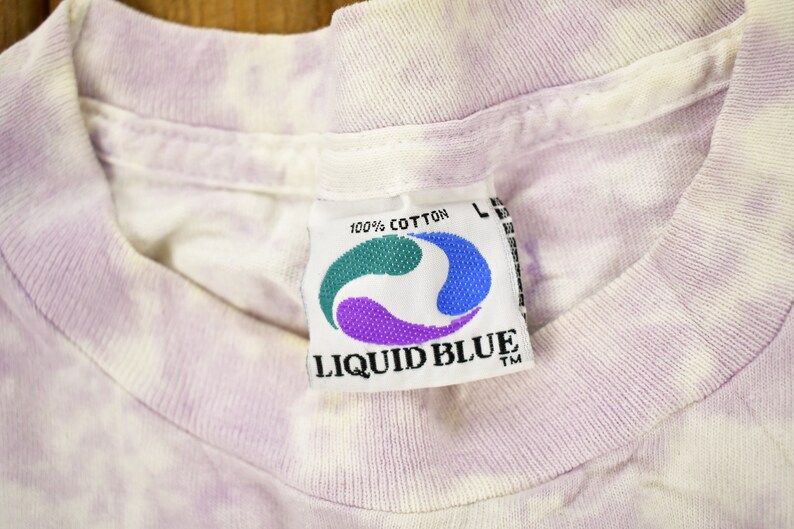 May include: A white and purple tie-dye t-shirt with a Liquid Blue tag. The tag is white with a blue and purple swirl design and the text "100% Cotton" and "Liquid Blue TM".