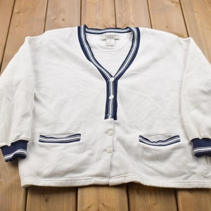 May include: White cardigan with blue and white striped trim around the neckline, cuffs, and pockets. The cardigan has a button closure and is made of a soft, comfortable fabric.