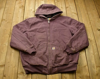 Puede incluir: Una chaqueta Carhartt con capucha de color morado, con cierre de cremallera y un pequeño parche con el logotipo de Carhartt en el pecho izquierdo.