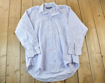 May include: Light blue button-down shirt with long sleeves. The shirt has a classic collar and a small embroidered logo on the chest. The fabric appears to be a lightweight cotton or blend. The shirt is laid flat on a wooden surface.