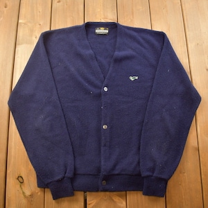 以下が含まれることがあります： 左胸に緑色の刺繍ロゴが付いた紺色のカーディガン。Vネックでボタン留めです。