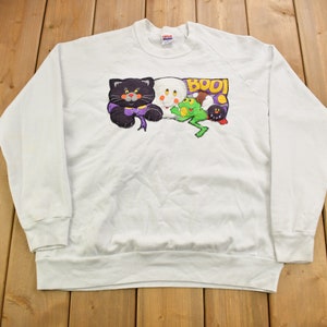 Puede incluir: Sudadera blanca con un gráfico de temática de Halloween que presenta un gato negro, un fantasma, una rana y la palabra "BOO!"
