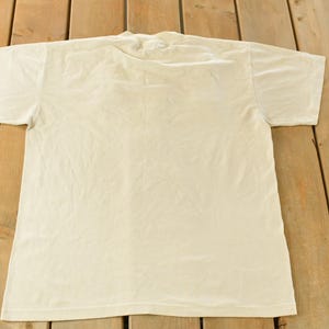 K&ouml;nnte beinhalten: Ein schlichtes, cremefarbenes T-Shirt mit kurzen &Auml;rmeln, flach auf einer Holzoberfl&auml;che liegend. Das Shirt besteht aus einem weichen Material und hat ein schlichtes, klassisches Design. Ein vielseitiger Kleiderschrank-Klassiker.