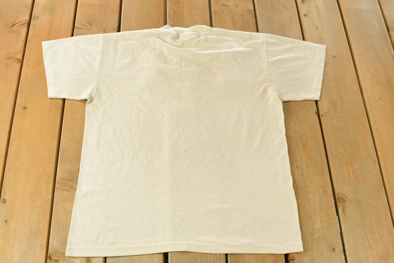 K&ouml;nnte beinhalten: Ein schlichtes, cremefarbenes T-Shirt mit kurzen &Auml;rmeln, flach auf einer Holzoberfl&auml;che liegend. Das Shirt besteht aus einem weichen Material und hat ein schlichtes, klassisches Design. Ein vielseitiger Kleiderschrank-Klassiker.
