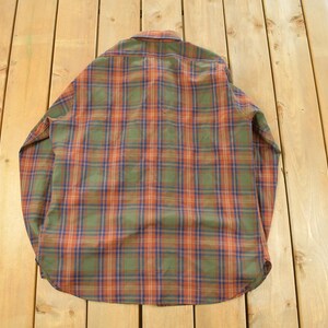 May include: A vintage button-down shirt with a classic plaid pattern in shades of green, orange, and blue. The long-sleeved shirt is folded and lies on a wooden surface.
