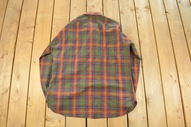 May include: A vintage button-down shirt with a classic plaid pattern in shades of green, orange, and blue. The long-sleeved shirt is folded and lies on a wooden surface.