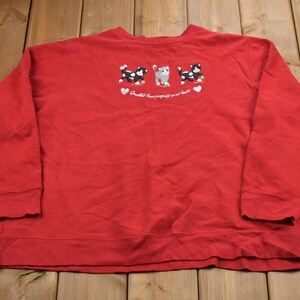 Puede incluir: Sudadera roja de manga larga con un gráfico de tres gatos blanco y negro con corazones rojos. El texto "Pawsitive thoughts are our best bet" está bordado debajo de los gatos.