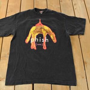 May include: Black t-shirt featuring a graphic design with the word "phish" in white, and the word "hoist" in parentheses. The graphic is a yellow and orange image. The t-shirt is made of a soft material.