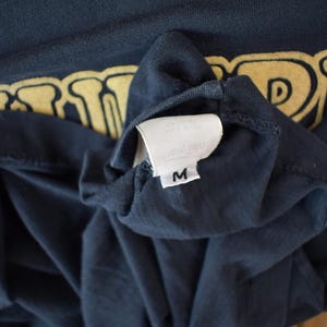 May include: A dark blue t-shirt with a yellow graphic print of the word "DRAGON". The shirt has a white tag with the letter "M" on it.