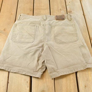 May include: Khaki-colored shorts with multiple pockets, including two back pockets and a side pocket. The shorts have a brown leather patch on the back waistband. The shorts are made of a durable fabric.