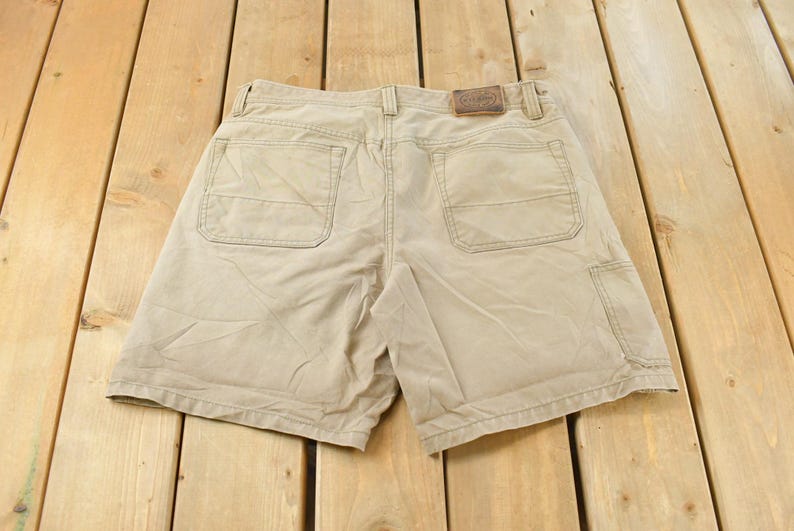 May include: Khaki-colored shorts with multiple pockets, including two back pockets and a side pocket. The shorts have a brown leather patch on the back waistband. The shorts are made of a durable fabric.