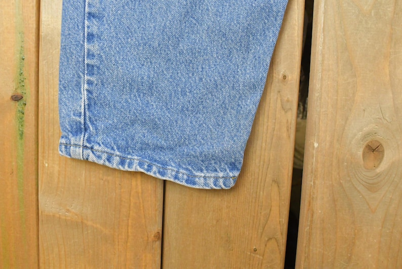 Puede incluir: Primer plano de una pernera de vaqueros azul claro. El dobladillo es visible, mostrando las costuras y el borde de la tela. Los vaqueros est&aacute;n sobre una superficie de madera.