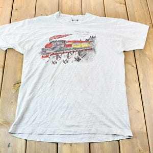 May include: A light grey t-shirt featuring a vintage-style illustration of a train travelling through a snowy forest. The text on the shirt reads "Canadian & Western Railroad - Calgary".