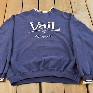 May include: Navy blue sweatshirt with the word "Vail" in white script across the chest, along with the word "Colorado" below. The sweatshirt has a crew neck with white trim and ribbed cuffs with white stripes.