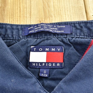 Pode incluir: Uma camisa azul marinho com colarinho e um patch com o logotipo da Tommy Hilfiger em vermelho e branco. A camisa &eacute; feita de 100% algod&atilde;o e tem uma etiqueta de tamanho que diz "S P/CH".