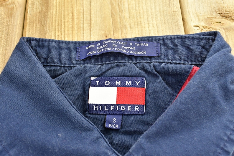 Pode incluir: Uma camisa azul marinho com colarinho e um patch com o logotipo da Tommy Hilfiger em vermelho e branco. A camisa &eacute; feita de 100% algod&atilde;o e tem uma etiqueta de tamanho que diz "S P/CH".