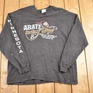 May include: A charcoal gray long-sleeve shirt featuring the text "MINNESOTA" on the left sleeve. The front displays a graphic with the text "ABATE STATE RALLY 8th ANNUAL" and event details. The shirt is likely vintage.