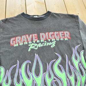 May include: A dark gray long-sleeve shirt featuring the text "GRAVE DIGGER MONSTER TRUCK Racing" in red and green. The shirt has a flame design in green and purple along the bottom.