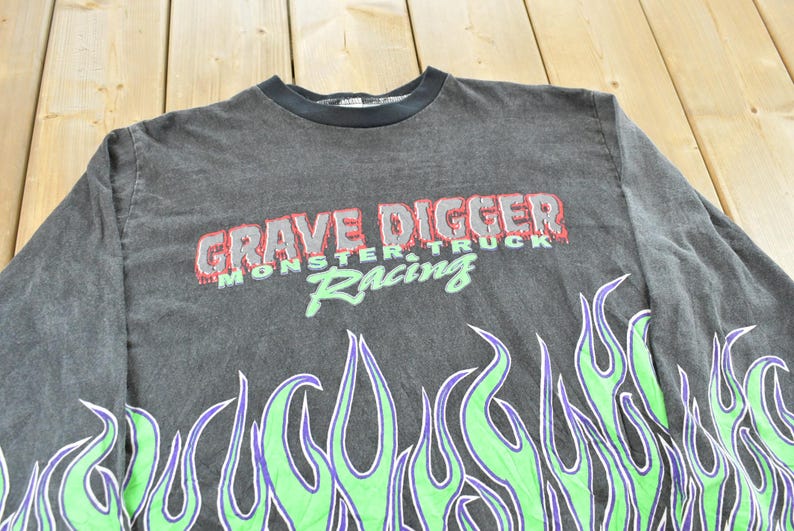 May include: A dark gray long-sleeve shirt featuring the text "GRAVE DIGGER MONSTER TRUCK Racing" in red and green. The shirt has a flame design in green and purple along the bottom.