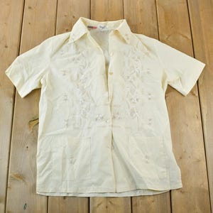 May include: Cream-coloured short-sleeved button-down shirt with floral embroidery. The shirt has a collared neckline and a single pocket. The garment is laid flat on a wooden surface.