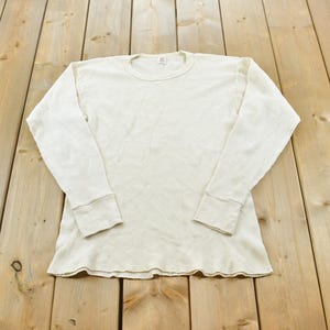 May include: A long-sleeve thermal shirt in an off-white colour featuring a textured waffle-knit pattern, a crew neckline, and ribbed cuffs at the wrists. The garment is constructed with a standard fit and a straight hemline at the bottom edge.