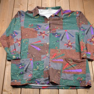 May include: A long-sleeved button-up shirt with a colourful geometric pattern. The shirt features a variety of colours, including green, brown, purple, and blue. The pattern includes abstract shapes, stars, and crescent moons.